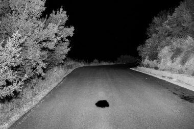 A black and white photograph of a road in Crete. There is a black hole in the road.