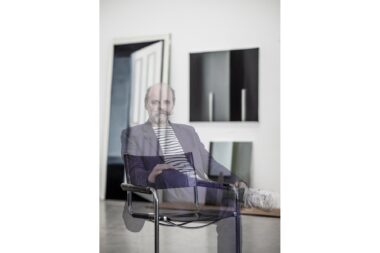 A multiple exposure portrait of the artist Gavin Turk with his paintings of candles in the background.