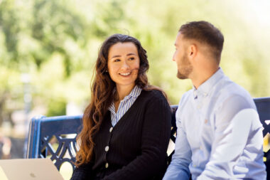 A lifestyle photograph of two people talking on a bench in a park.