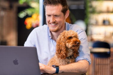 A man working at a laptop in a cafe in London with a dog sat beside him.
