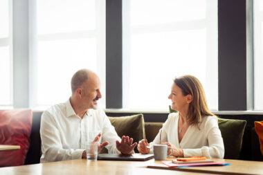 Two business professionals laughing in casual meeting by window, man gesturing while talking with laptop and coffee on table- corporate lifestyle photography.
