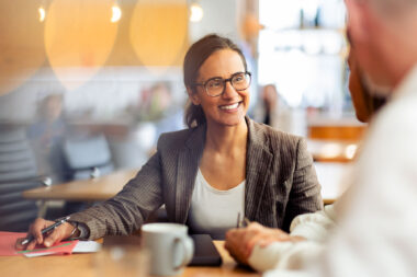 Business meeting stock photo: smiling businesswoman in blazer taking notes with colleague at coffee shop table, corporate lifestyle photography