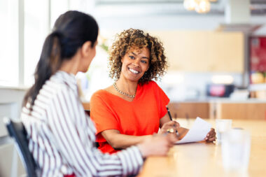Smiling businesswoman with in orange top holding paperwork, talking with colleague in office meeting, contemporary corporate lifestyle photography.