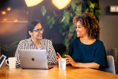 Two businesswomen smiling and chatting at desk with Apple MacBook laptop, warm office setting with plants and string lights, modrn corporate lifestyle photography.