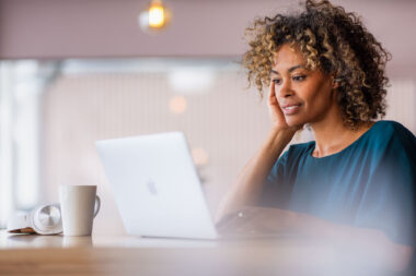 Woman relaxing at desk looking at Apple MacBook laptop, coffee mug and headphones nearby — lifestyle photography.